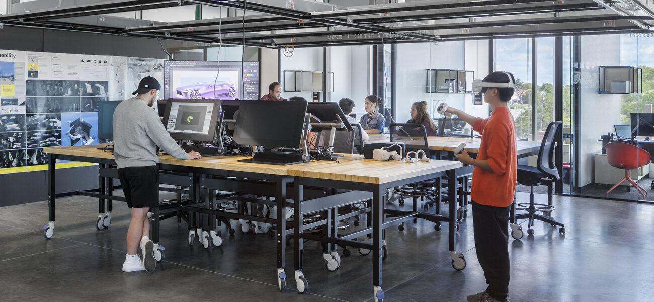 One man stands working at a computer while another, wearing a virtual reality headset, moves devices with his hands.