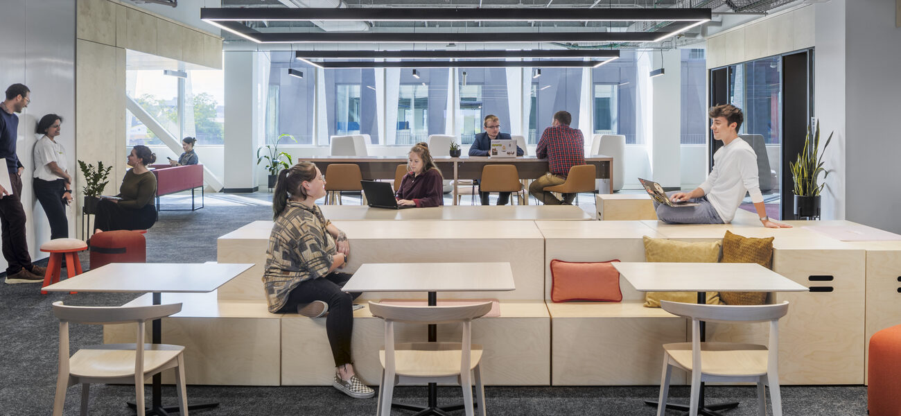 A group of adults sitting and standing, some working on their laptops while others chat.
