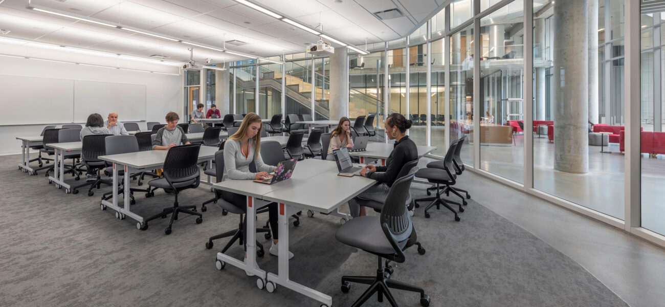 Students seated in a glass-fronted room where all of the tables and chairs are on wheels.