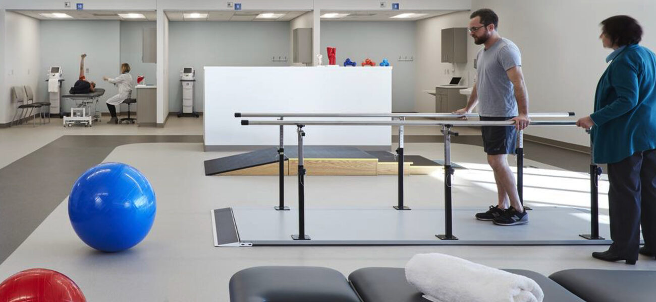 A man walks between two parallel handrails as a physical therapist looks on.