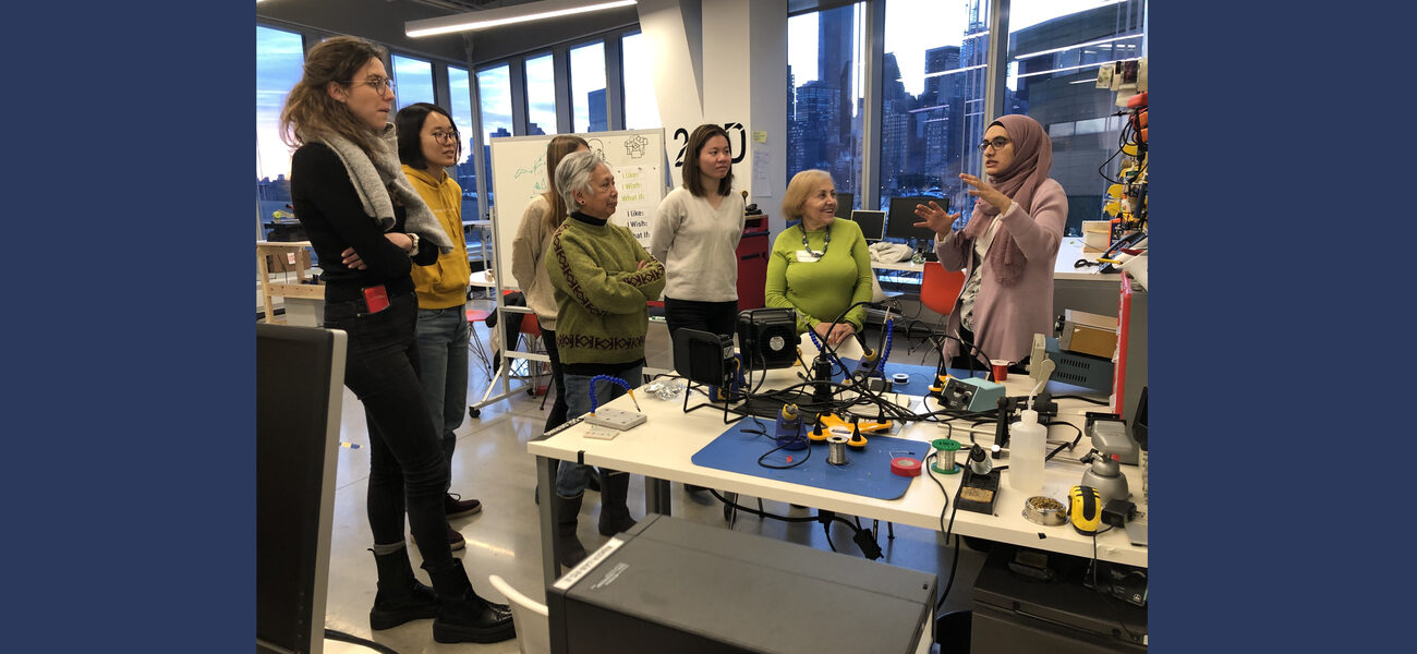 Five women and a man stand at a table coveredin tools as another woman describes the room.