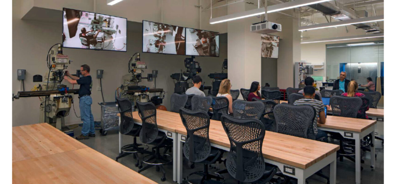 College students sitting at tables in a large classroom. Display screen hang on the walls beside them.