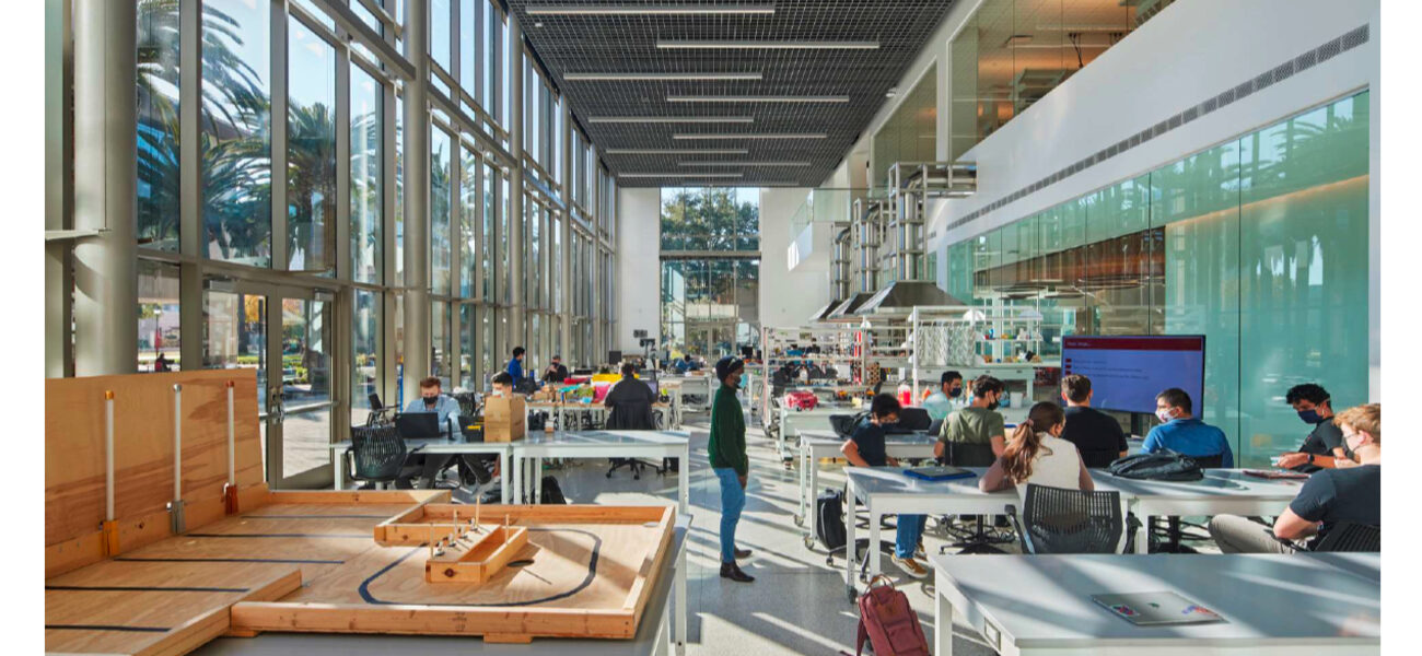 College students sit at large tables while other work with tools in an engineering project lab.