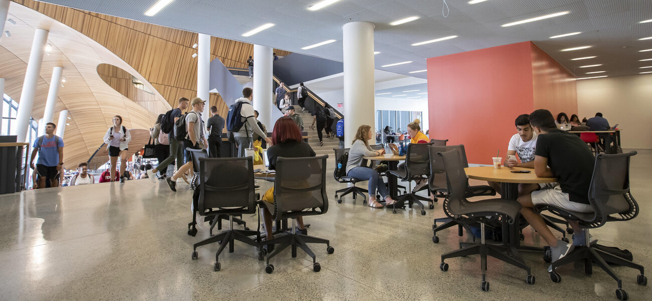 Students sit at multiple small tables in a landing between two large staircases. 