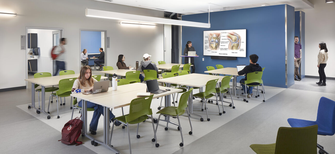 Students work at large tables. Anatomy diagrams are on the wall.