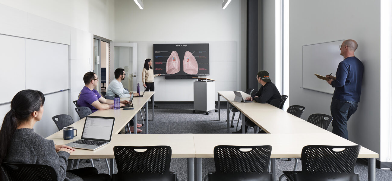 Students sit at a horseshoe-shaped table as a professor points to an anatomyy diagram on the wall