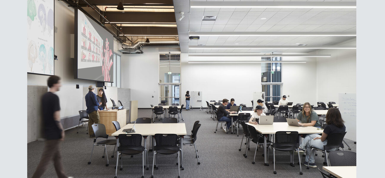 Students sit at multiple tables in front of several large video screens