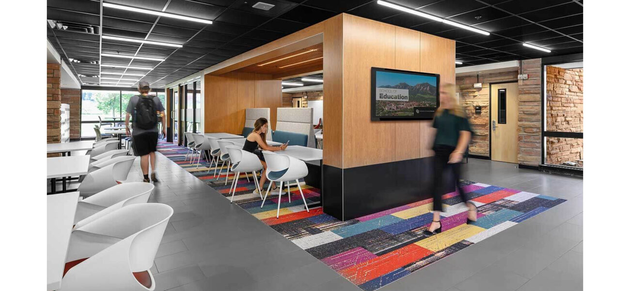 A woman works at a table in the center of the wide hallway, as another woman walks across brightly-colored floor tiles in the center of the space and a man walks toward a wall of windows to the outside..