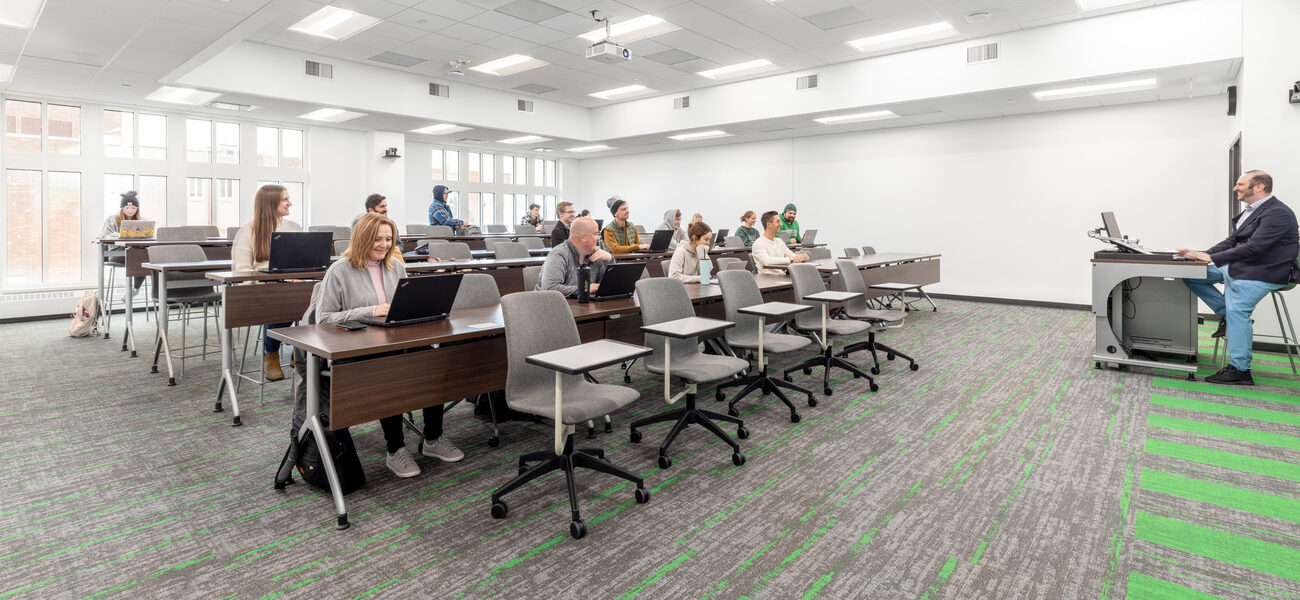 College students work on laptops at a row of desks in a large classroom.