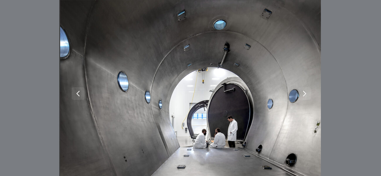 Thre men in lab coats sit at the entrance to a very large steel chamber with a flat floor and a rounded roof.