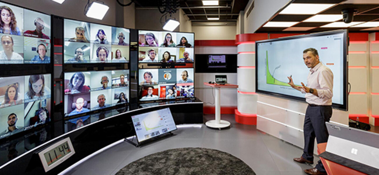 A man gestures in front of a wall of comptuer monitors showing the faces of his students.