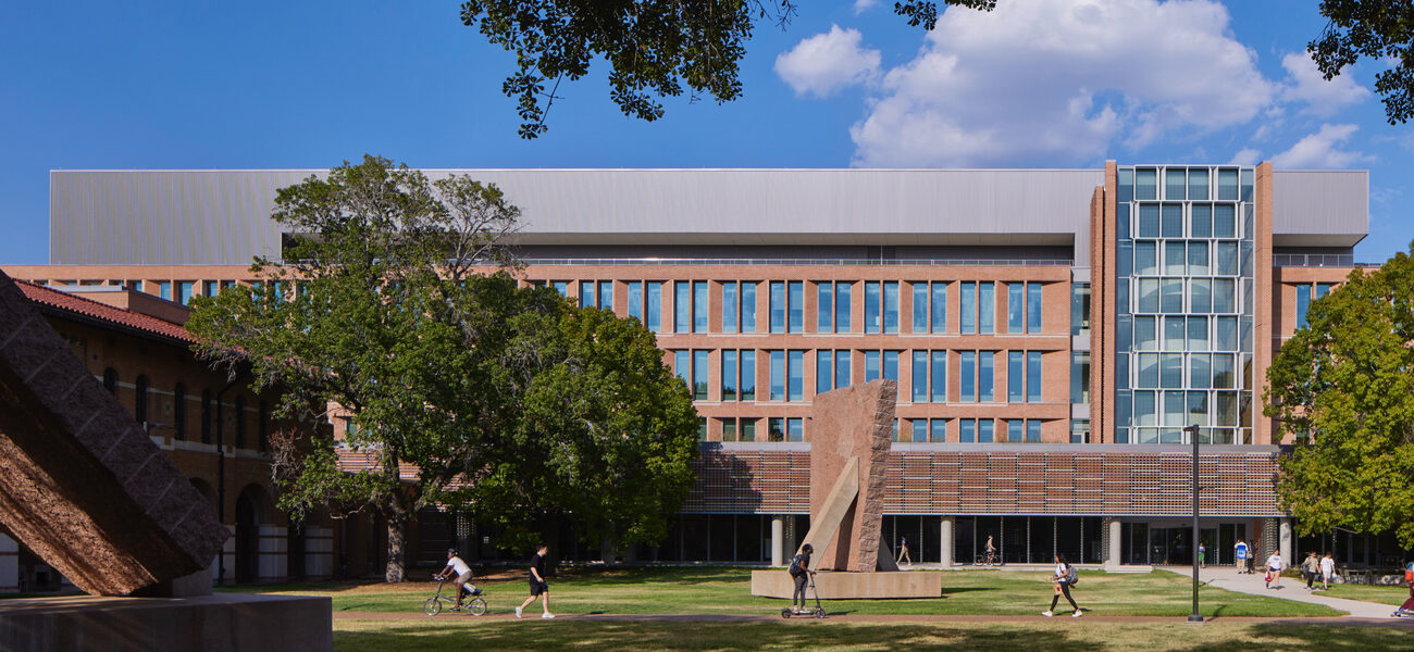 Long building with many sall windows, with a lawn in front where people are walking