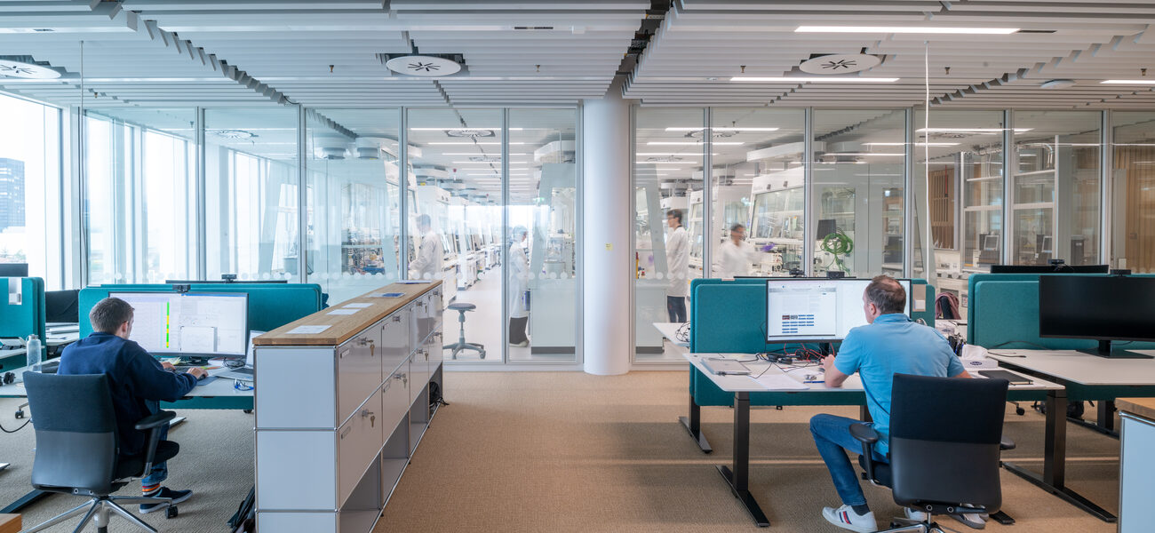 People work at compters in front of a wall of glass; on the other side of the glass is a lab.