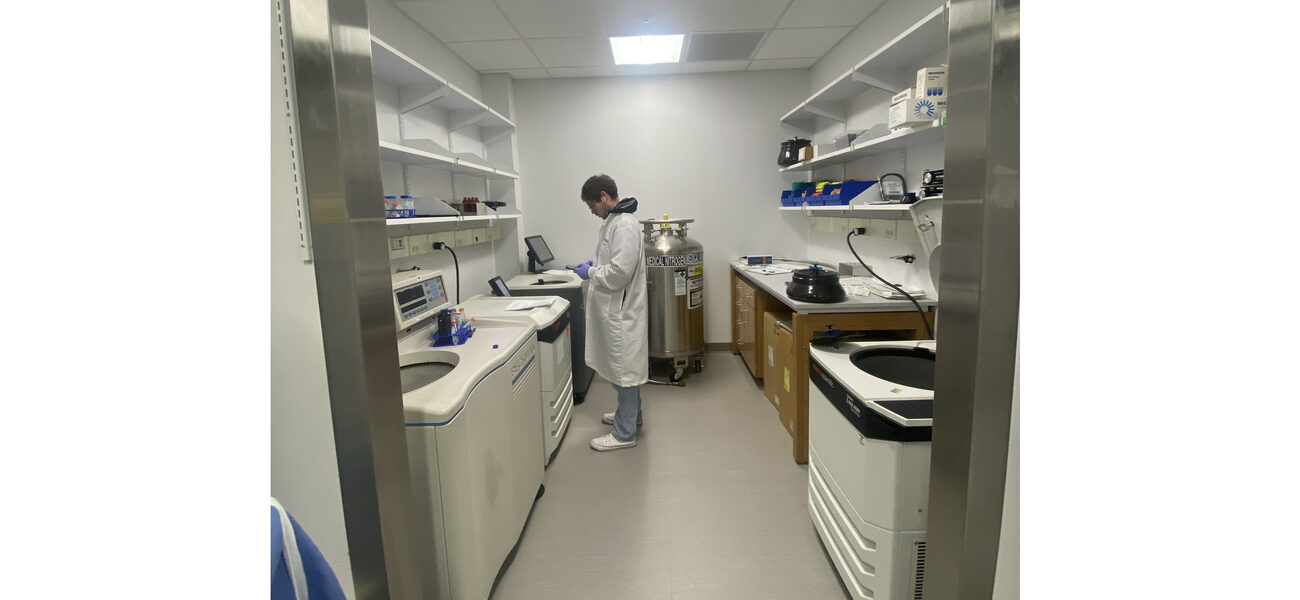A man in a long white lab coat works at an instrument in a small narrow room.