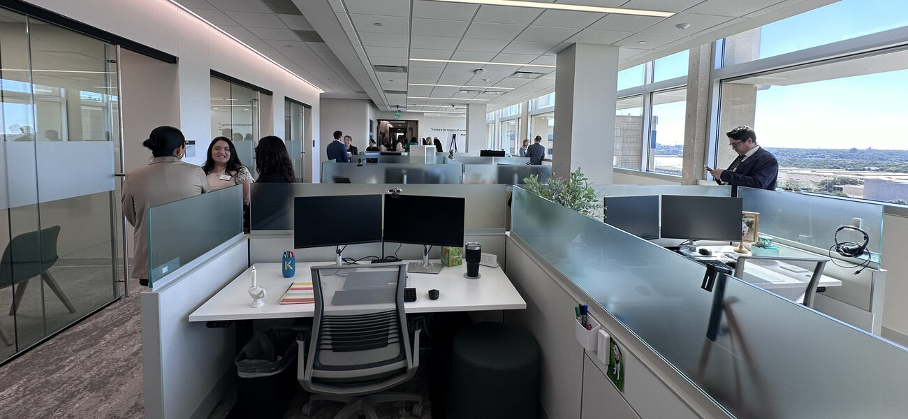 Three people stand chatting next to a small cubicle, in a room full of cubicles.