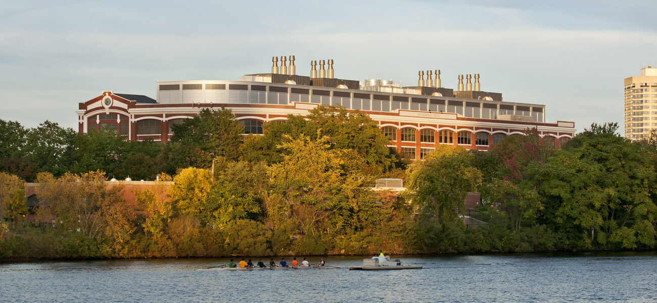 Long brick building topped with exhaust stacks, in front of a river with boaters on it.