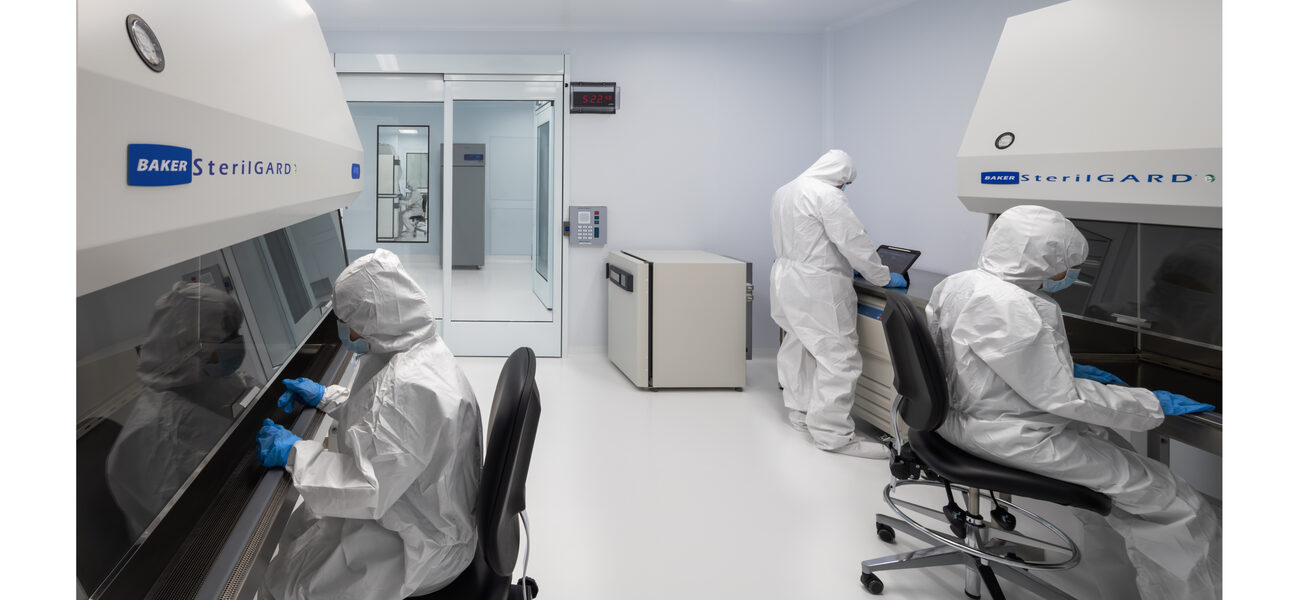 three people in head-to-foot protective suits -- two seated, one standing -- work in a lab. 