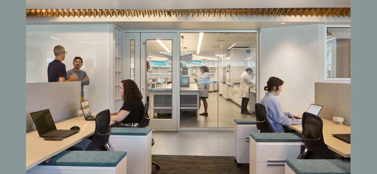 Two women sit at long benches working on laptops. Across the hall is a lab.