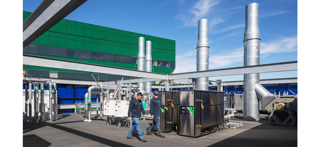two men walk on a flat roof past large pieces of machinery