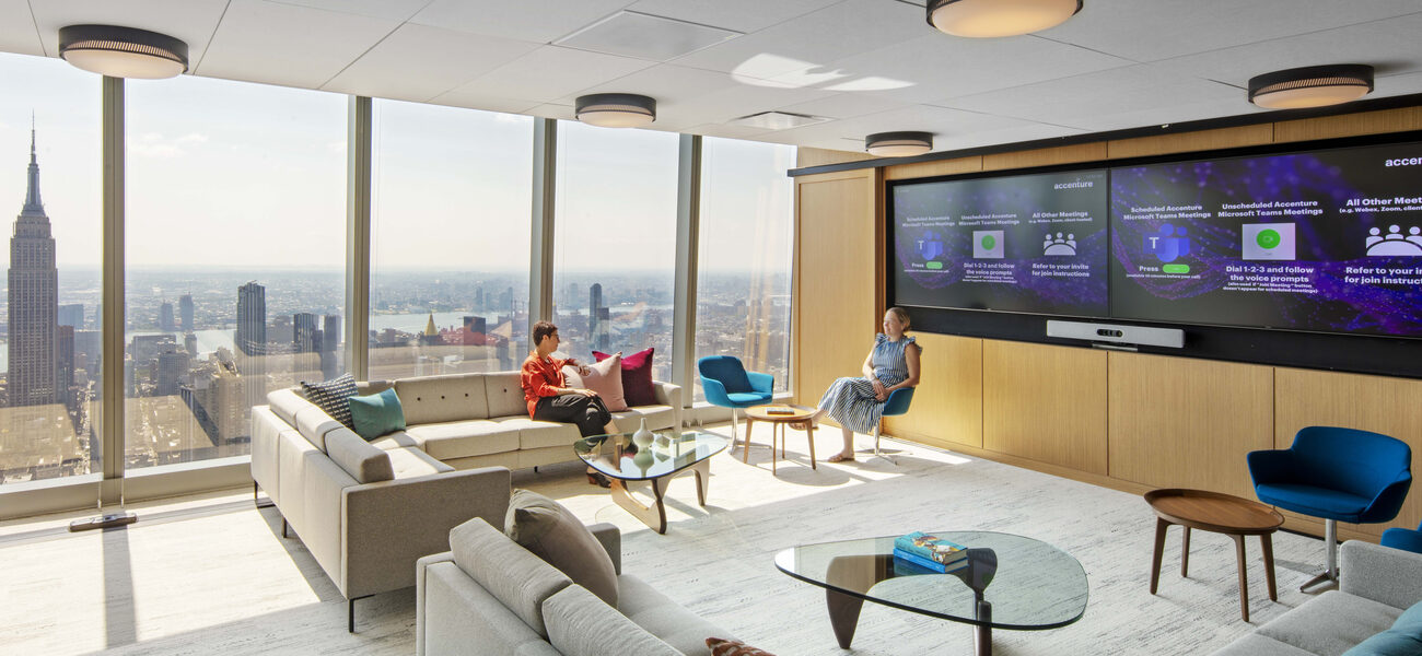 Two people sit in a room with a wall of computer screens and several couches and chairs