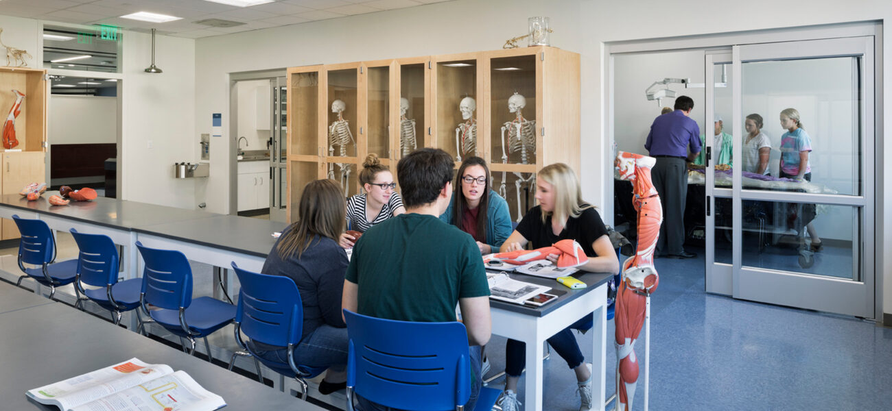 A man and three women sit at the end of a long table; behind them, another group works in a small room behind a glass door.