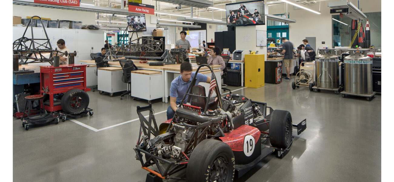 In a large room full of mechanical equipment, a man works on a vehicle.