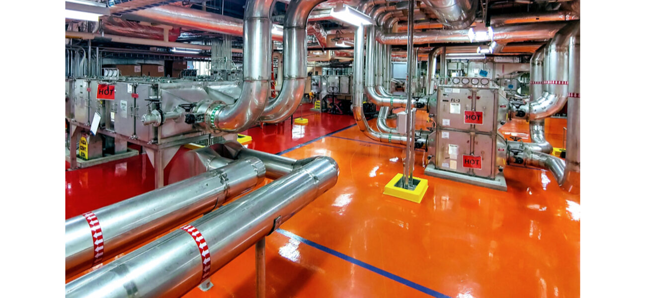 A tangle of metal pipes runs along the floor and ceiling of a large open space, attched to pieces of mechanical equipment.