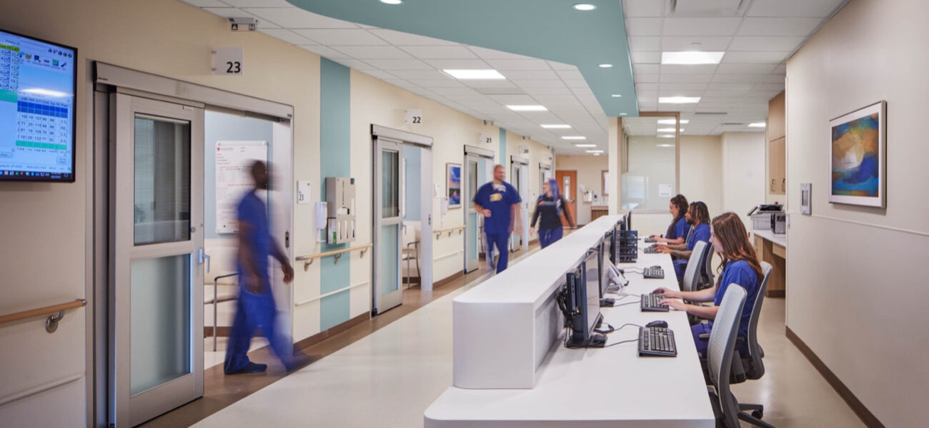 Two women and a man sit at a long desk working on computers, while three people pass by in the hallway in front of them.