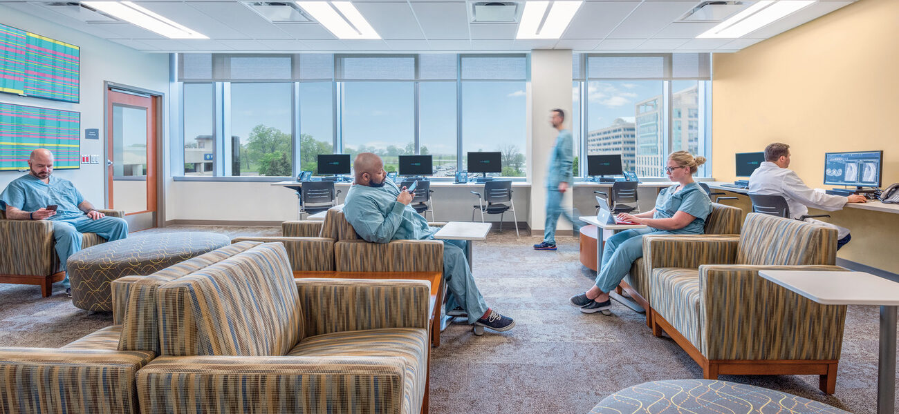 Two men and a woman sit on upholstered chairs while another man sits on a desk chair working on a laptop in a room with a wall of windows.