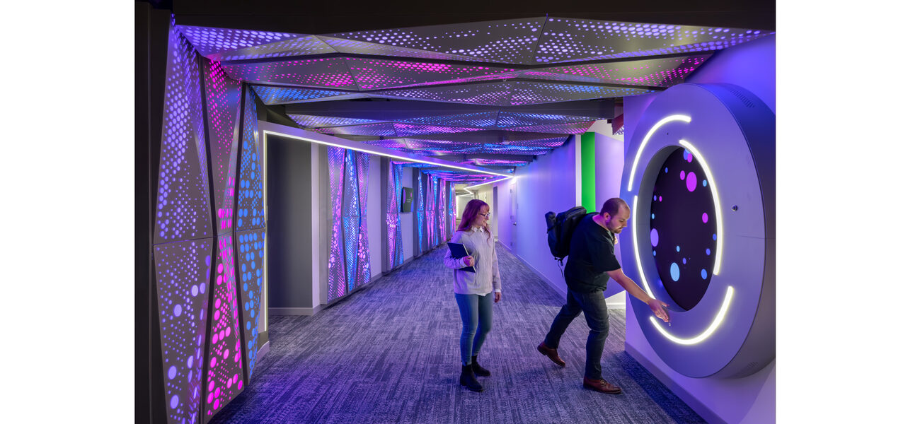 A man controls a floor-to-ceiling computer touch-screen on a wall in a long hallway; a woman stands behind him.