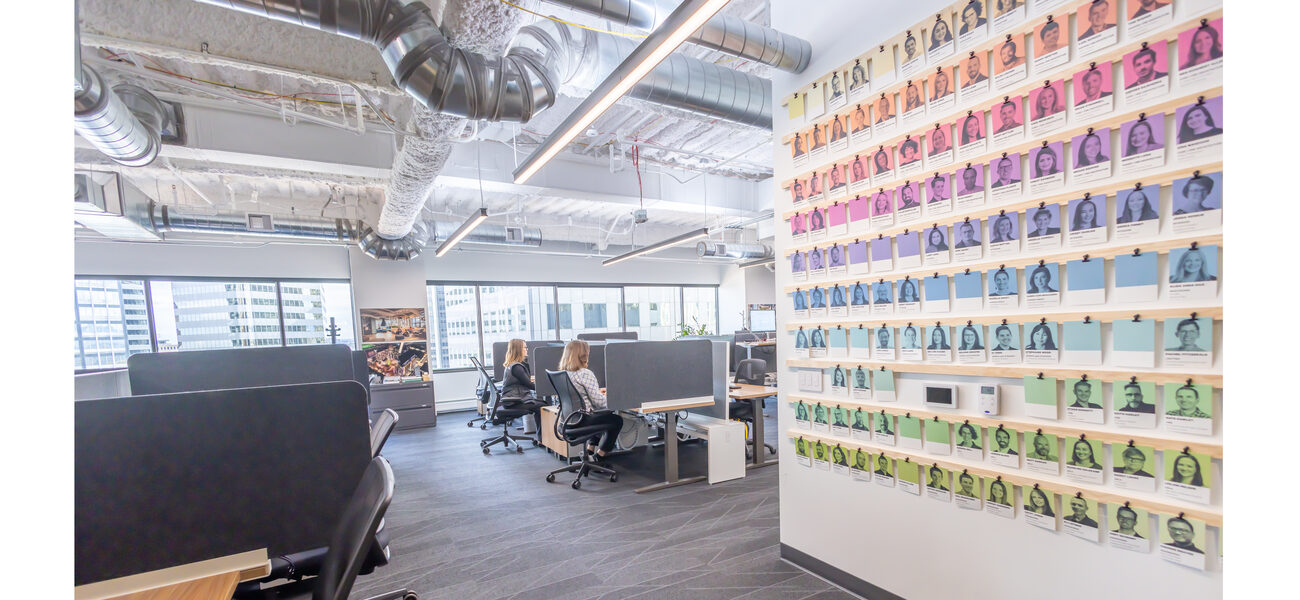 Two women sit at desks behind a large wall covered in photos of men and women.