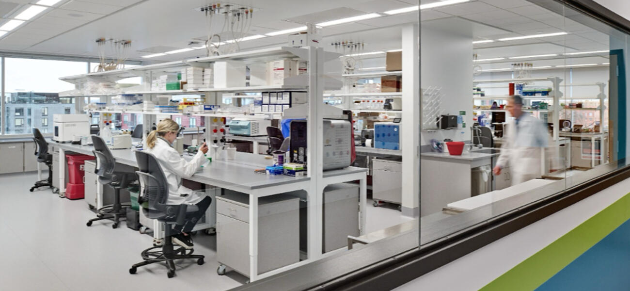 A woman sits in a chair working at a lab bench while a mn walks by.