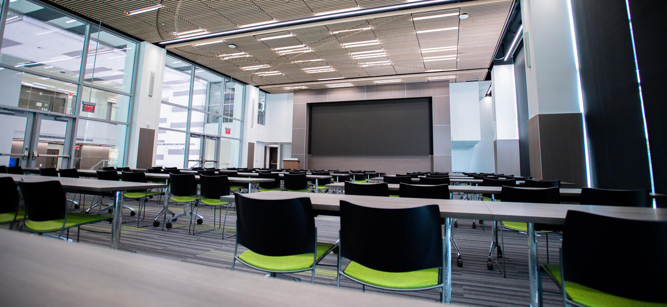 Rows of charis at rows of tables facing a screen that covers an entire wall.