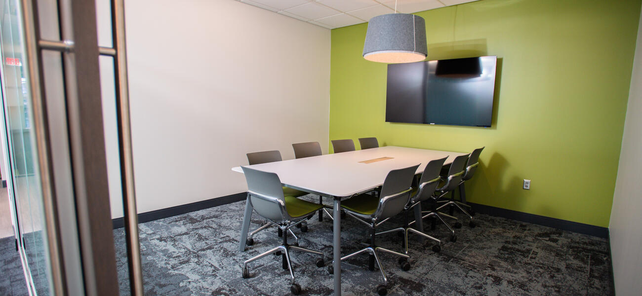 Nine chairs sit around a rectangular table in front of a large computer monitor hanging on the wall.