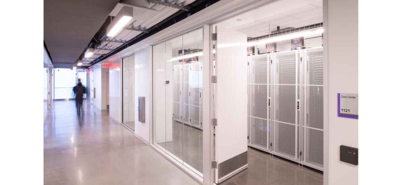 A stark, brightly-lit hallway lined with glass behind which are massive banks of computers