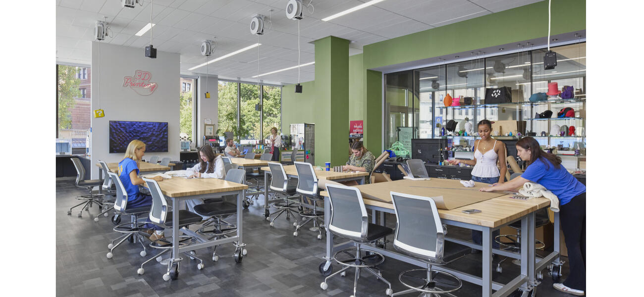 Men and women sit and stand, working at tables in a large open room.