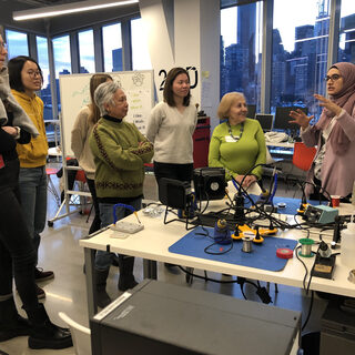 Five women and a man stand at a table coveredin tools as another woman describes the room.