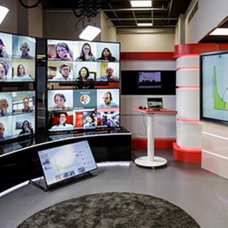 A man gestures in front of a wall of comptuer monitors showing the faces of his students.