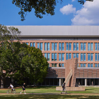 Long building with many sall windows, with a lawn in front where people are walking