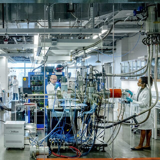Two women and one man work on lab benches in a room full of electronic equipment.