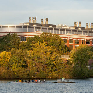 Long brick building topped with exhaust stacks, in front of a river with boaters on it.