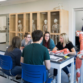 A man and three women sit at the end of a long table; behind them, another group works in a small room behind a glass door.