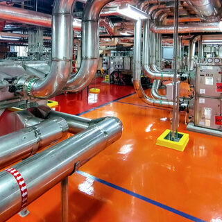 A tangle of metal pipes runs along the floor and ceiling of a large open space, attched to pieces of mechanical equipment.