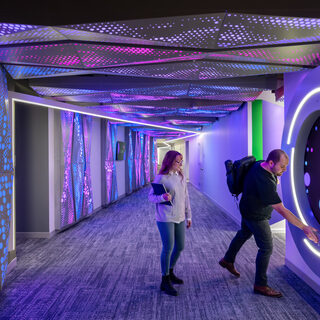A man controls a floor-to-ceiling computer touch-screen on a wall in a long hallway; a woman stands behind him.