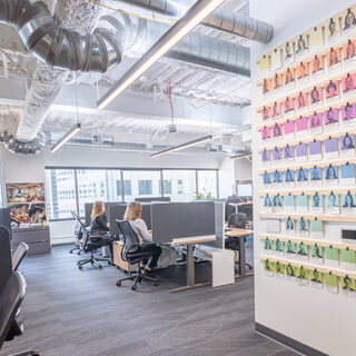 Two women sit at desks behind a large wall covered in photos of men and women.